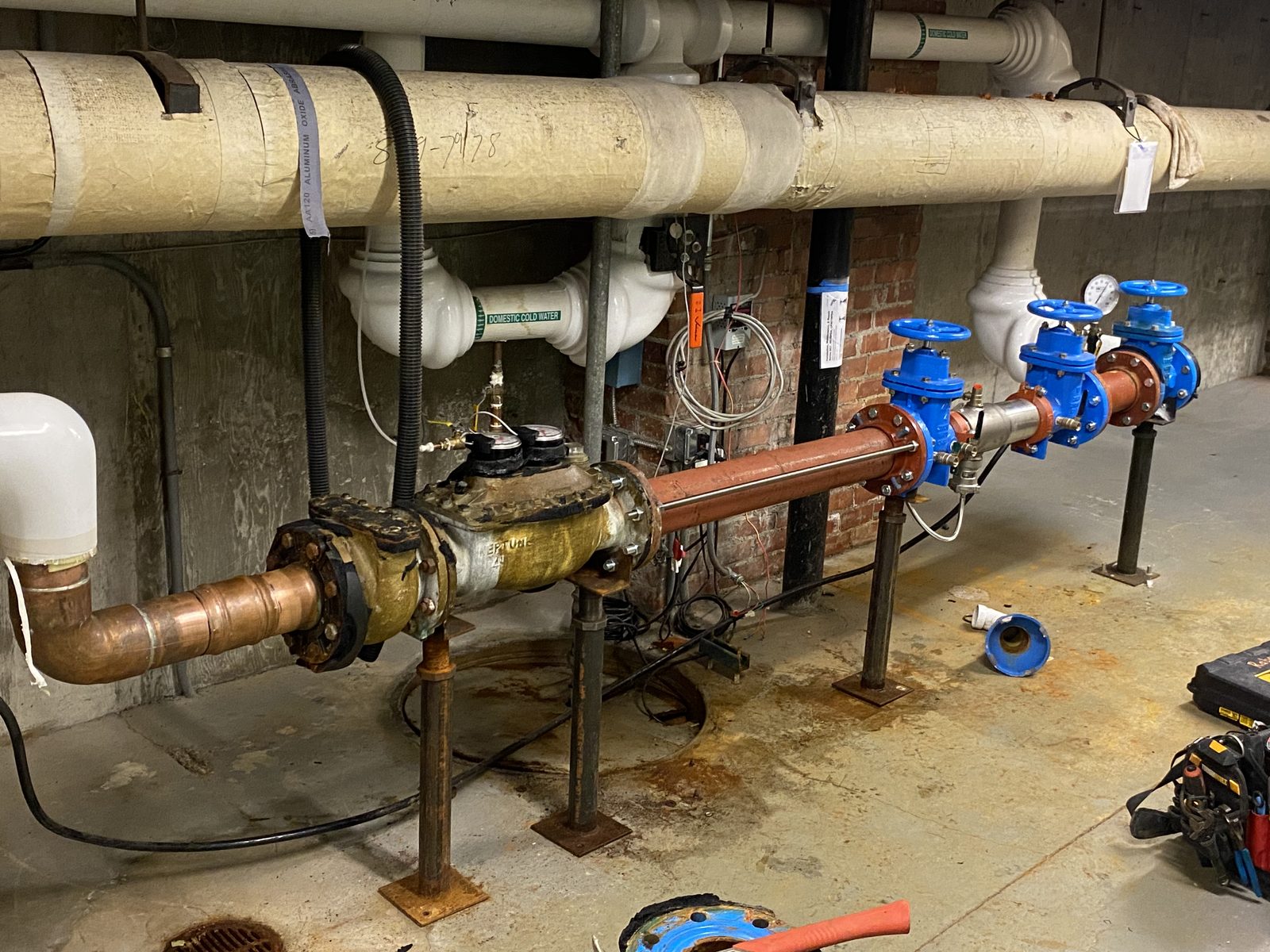 A long water meter and backflow assembly with blue gate valves on steel stands in an old brick basement