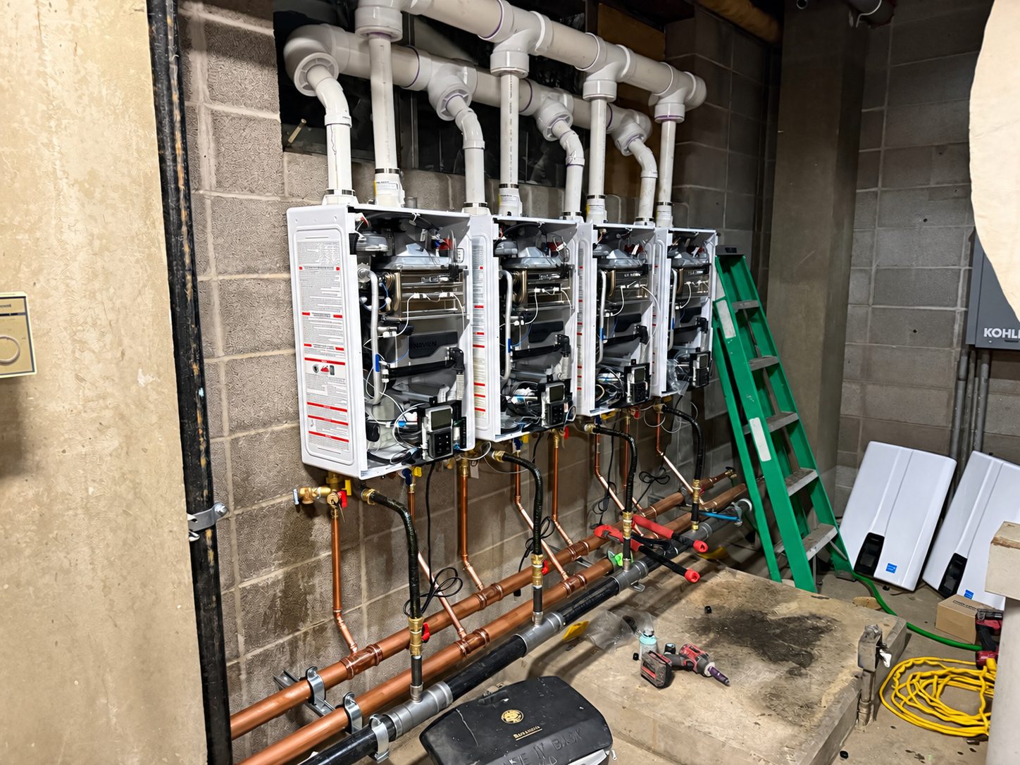 Four Navien tankless water heaters installed side by side on a block wall in a commercial basement, with a new copper manifold and white PVC venting